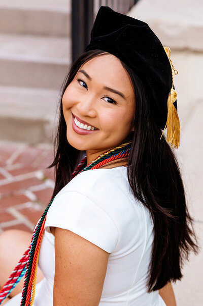 Masters student wearing cords and cap sitting on steps smiling at the camera
