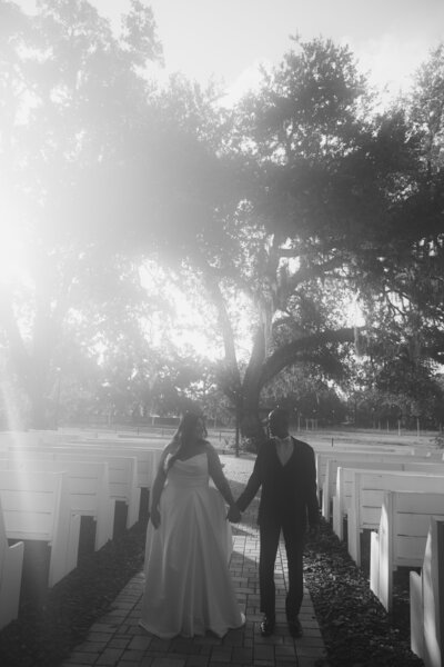 A black and white shot of a newlywed couple walking away from the camera down a paved path lined with white markers. The bride wears a long gown and the groom is in a dark suit, holding hands as they walk toward large trees in the distance.