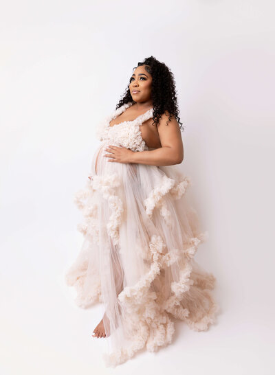Expectant mom in fitted black grown poses for maternity photos in Brooklyn, NY photo studio. She has one hand on her back and one hand under her bump. She is looking up at the light and smiling.