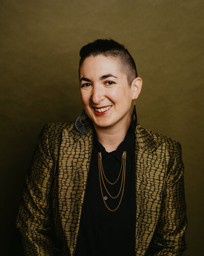 A woman with short, dark hair and a unique undercut smiles at the camera. She is wearing a green textured jacket with gold chains, and the background is a muted green, giving the image a stylish and edgy vibe.