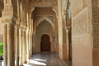 A hallway with intricately carved arches and columns, showcasing detailed Arabic patterns and designs. Sunlight filters through, highlighting the ornate details. A dark wooden door is visible at the end of the corridor, reminiscent of a personalized travel planning journey to hidden European gems.