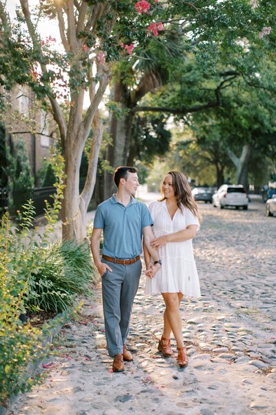 Smiling man and woman holding hands while walking down a cobblestone street in Charleston South Carolina with the morning sun peeking through the trees behind them.