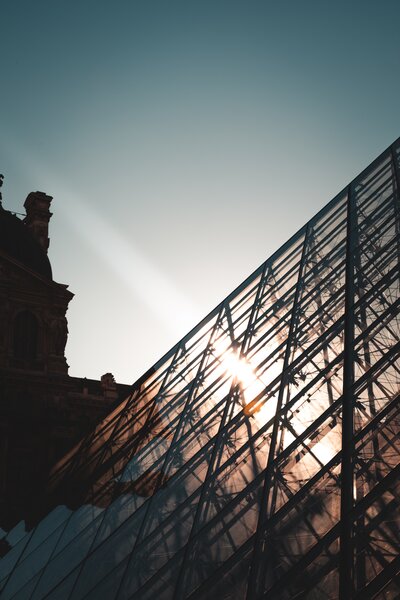 A photograph of a section of the Louvre's glass pyramid with the sun setting behind and the silouette of a nearby building.