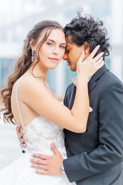 A bride holds her grooms face as she looks at the camera and presses his face against hers. They are standing on a rooftop of a hotel.