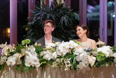 A newlywed couple smiling and sitting at a table covered in florals 