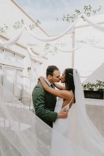 A bride and groom share a kiss beneath soft white draping and greenery at SoHo63, a Chandler wedding and event venue. Natural light fills the space as the couple embraces in front of the fireplace, with the bride’s veil gently trailing behind her.