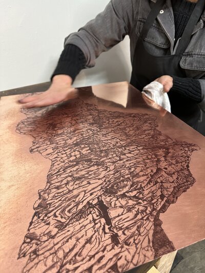 A printmaker carefully wipes a large, intricately etched copper plate, revealing the detailed artwork for a print.