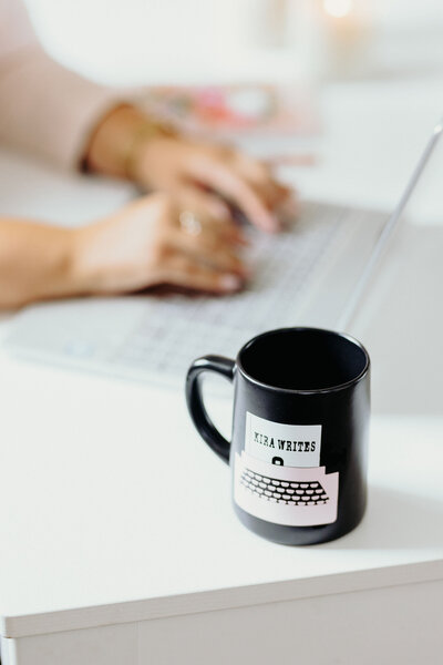 Kira Writes branded mug is in focus while Kira is typing on her computer in the background.