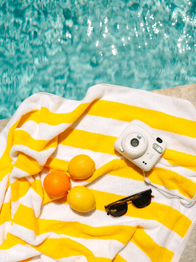 Polaroid camera, sunglasses, and lemons and oranges laying on a yellow striped towel next to a pool