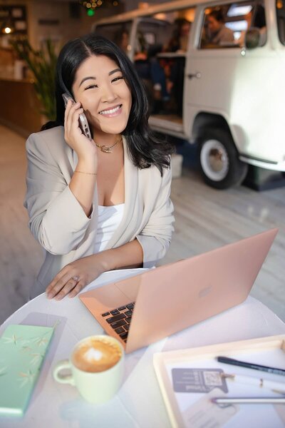 Smiling woman in a light blazer talks on the phone while working on a rose gold laptop at a café table with coffee and notebooks. Captured during a Seattle personal branding session, this image conveys confidence, professionalism, and connection for modern women in business.