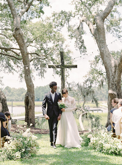 Charleston South Carolina wedding with all of the European feel. Bride and groom walking down aisle after ceremony. She has on a long sleeved lace gown and he is wearing a black tuxedo. She has a bouquet of baby's breath and white florals and greenery form the aisle walkway on either side. The guests are sitting on wooden chairs. Photographed at River Oaks by wedding photographers in Charleston Amy Mulder Photography.