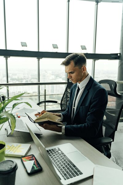A man in a navy suit sitting at a desk in a modern office, reading a notebook with laptops open.