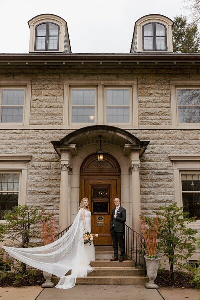 bride and groom stand outside on the steps of doorway with columns while someone holds bridal train out of frame in image by tom thornton st paul wedding photographer