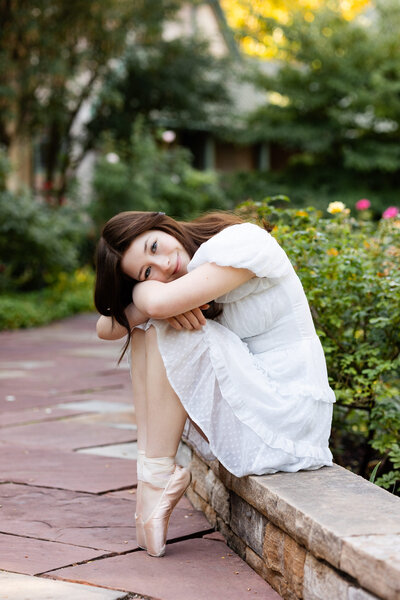 A ballerina sits on a ledge with her ballet shoes on and hugs and rests her head on her knees. 
