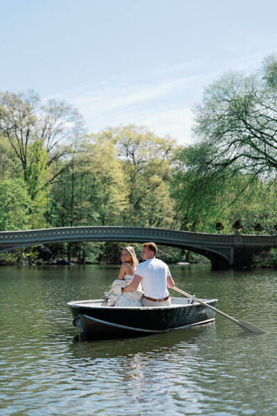 Couple romantically seated in a rowboat under the Bow Bridge in Central Park, New York City, surrounded by lush greenery and tranquil water.