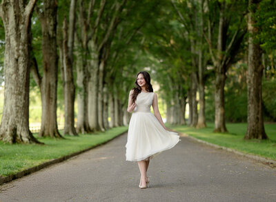 Brown haired girl with white dress walking on a street lines with trees and swishing her dress out. Photo taken by Jenni Nikki Photography at Berry College in Rome, GA (college outside of Atlanta, GA).