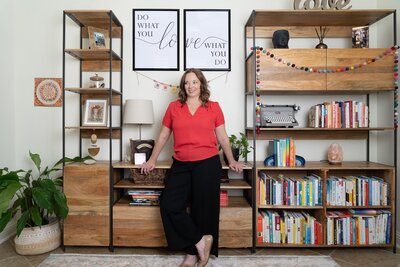 woman with curly brown hair wearing a red shirt and leaning against a bookshelf