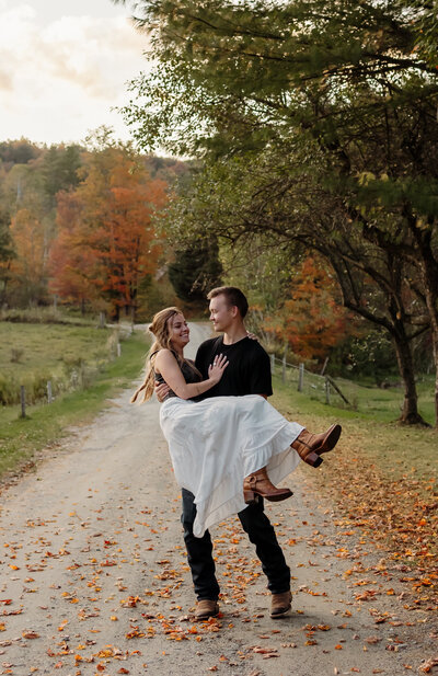 A fine art New England photographer documents an engagement photoshoot as the bride-to-be is picked up and spun around by her partner. They share a laughter while surrounded by the fall foliage of the New England trees—an intimate and timeless scene perfect for nature-loving couples.