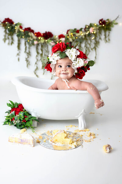 Baby looking off to the side, wearing a flower bonnet, surrounded by red flowers and hanging greenery with twinkle lights in the background.