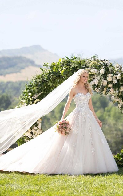 A modern approach to a classic silhouette, this ballgown with 3D flowers from Martina Liana radiates romance! The bodice of this gown is sheer, with layers of tulle and a sweetheart neckline adding to the romance. From the waist, the tulle skirt extends into a full ballgown, embellished with 3D flowers, sparkle tulle and sequins, creating an incredible sparkle and unique texture. A slight train, complete with lace hem, finishes the look. The back of this modern ballgown with 3D flowers zips up beneath crystal buttons, and this wedding dress is available in plus sizes.