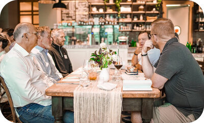 Group seated around a rustic wooden table in a restaurant, talking over drinks with a decorative runner and centerpiece.