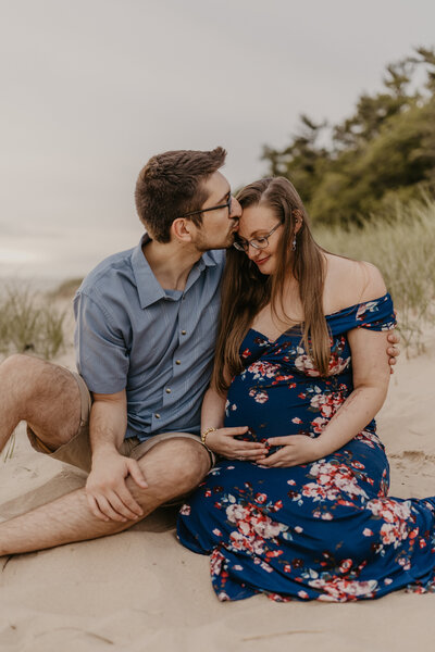 Couple sitting on Lake Michigan beach. The woman is holding her pregnant belly and man has his arms wrapped around her and is kissing her on the forehead.