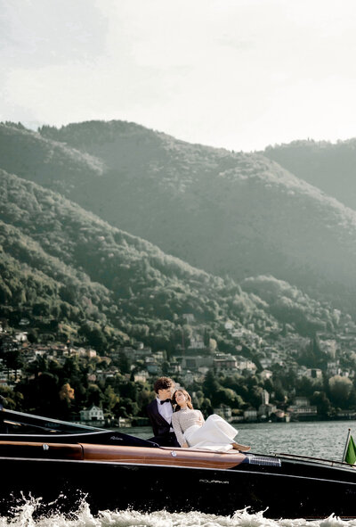 Newlywed couple in wedding attire cuddling on luxury wooden speedboat during scenic Lake Como elopement surrounded by mountains and Italian lakeside views