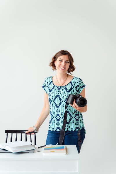 Calgary Brand Photographer Kate Daniel Photography, smiling, standing behind a desk, holding onto a black chair and her camera.