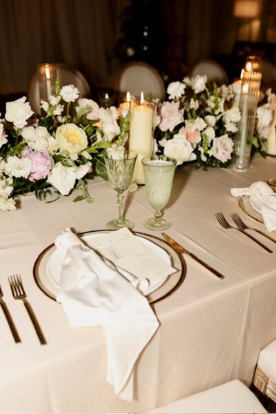 Elegant table setting with gold-rimmed plates, white napkins, vintage-style glasses, and gold cutlery, adorned with a floral centerpiece of white, pink, and yellow roses, and surrounded by glowing pillar candles.