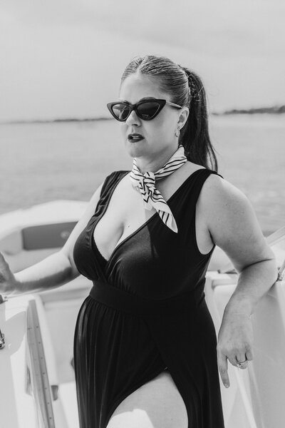 Black and white image of woman standing confidently on boat deck in flowing black dress and striped scarf, channeling vintage flair.