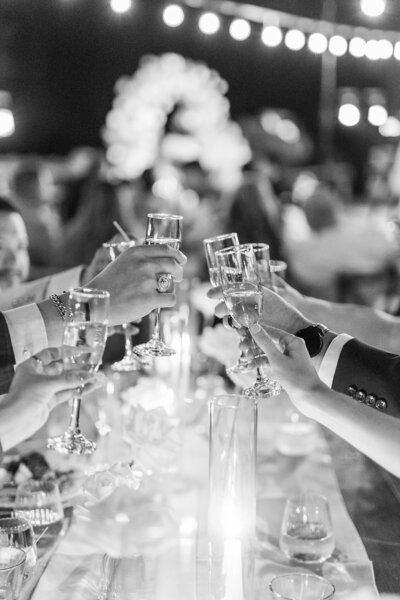 Black and white photo of wedding guests doing a champagne cheers during reception toasts, the candlelight glowing under the glasses, at Temecula Creek Inn.