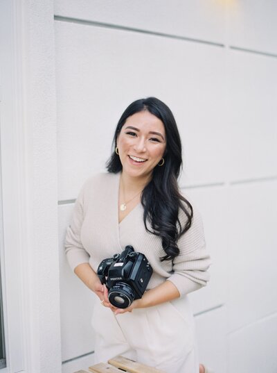 Texas fine art wedding photographer, Stephanie, holding her camera wearing a cream sweater and white dress pants