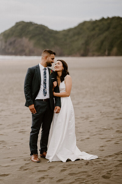 Couple who have just tied the know are standing together on Piha Beach for their bridal portraits