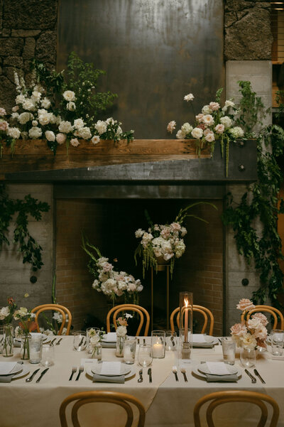 Wedding head table with natural bentwood chairs set in front of a fireplace, styled with simple, elegant decor.