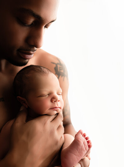 Father holding newborn baby son with skin on skin contact looking down at sun
