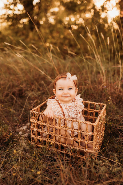 baby girl in a boho outfit sitting in a wooden basket in a field with tall grass