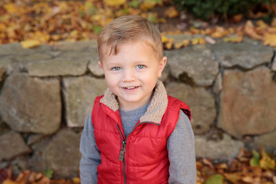 Little boy sits on log in yellow flowers with stuffed animal smiling at camera