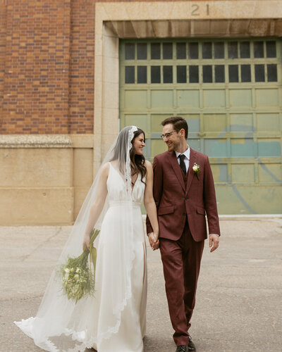 A bride and groom walk hand in hand outdoors in front of a grand building, embracing their newlywed journey.