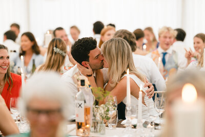 A West Morestead House wedding reception, a couple is kissing amidst smiling guests seated at tables. Candles and drinks add to the warm, festive atmosphere, making the historic venue even more enchanting.