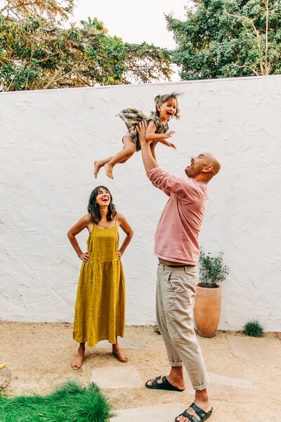 dad in pink shirt tossing toddler daughter in air while mom watches and smiles