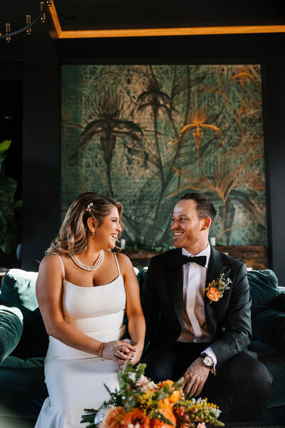 Bride and groom sitting on a couch holding hands and smiling at each other