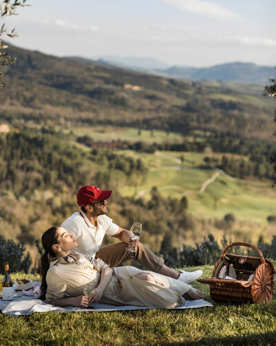 A couple relaxes on a picnic blanket on a grassy hill, overlooking a scenic landscape of rolling hills and valleys. They have a wicker basket and a bottle of wine. The man holds a glass, and both look serene under a clear sky.