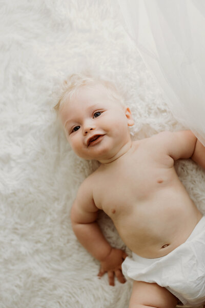 eight month old baby boy laying on his back on a white rug next to white sheer curtains in only a white diaper cover happily smiling and laughing at the camera