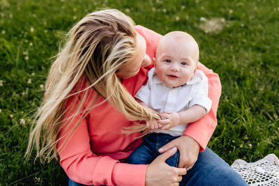 A mother sits on the grass holding her baby, who is reaching for her hair with a curious expression.