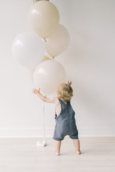 A toddler in a slate blue/gray romper reaching up toward a cluster of cream balloons in a light-filled studio — Portrait photos in Raleigh.