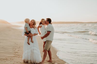Family of 4 plays on the brach during a family photo session. Mom is wearing a long white dress and is holding  her young son. Dad holds big sister and kisses her on the cheek