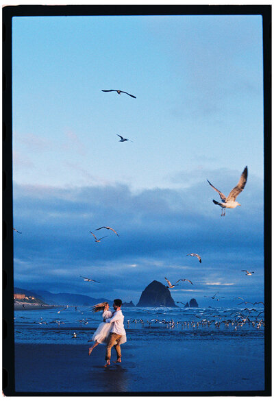 Couple dances barefoot on Cannon Beach at sunset with seagulls flying around them and Haystack Rock in the background; dreamy film tones and motion captured on analog photography.