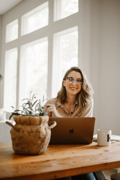 Smiling woman wearing glasses sitting at a dining table with laptop and mug