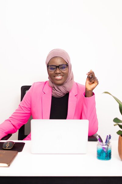 Photo of a woman in an office setting, sitting at a desk with a white laptop in front of her. She is smiling and wearing black-framed glasses. She is wearing a taupe headwrap, and wearing a pink blazer with a black shirt underneath.