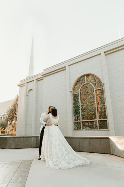 Photo taken by Bailee Freeman Photography of a bride and groom in front of the stained glass at the Boise Temple for the Church of Jesus Christ of Latter-day Saints. Groom is embracing the bride and showcasing the back of her dress.